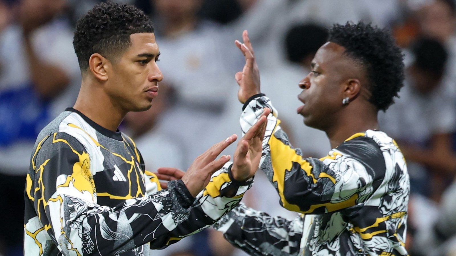 Vinícius Jr. et Jude Bellingham partagent un moment d'encouragement, portant tous deux des maillots à motifs frappants, dans l'atmosphère vibrante d'un stade.