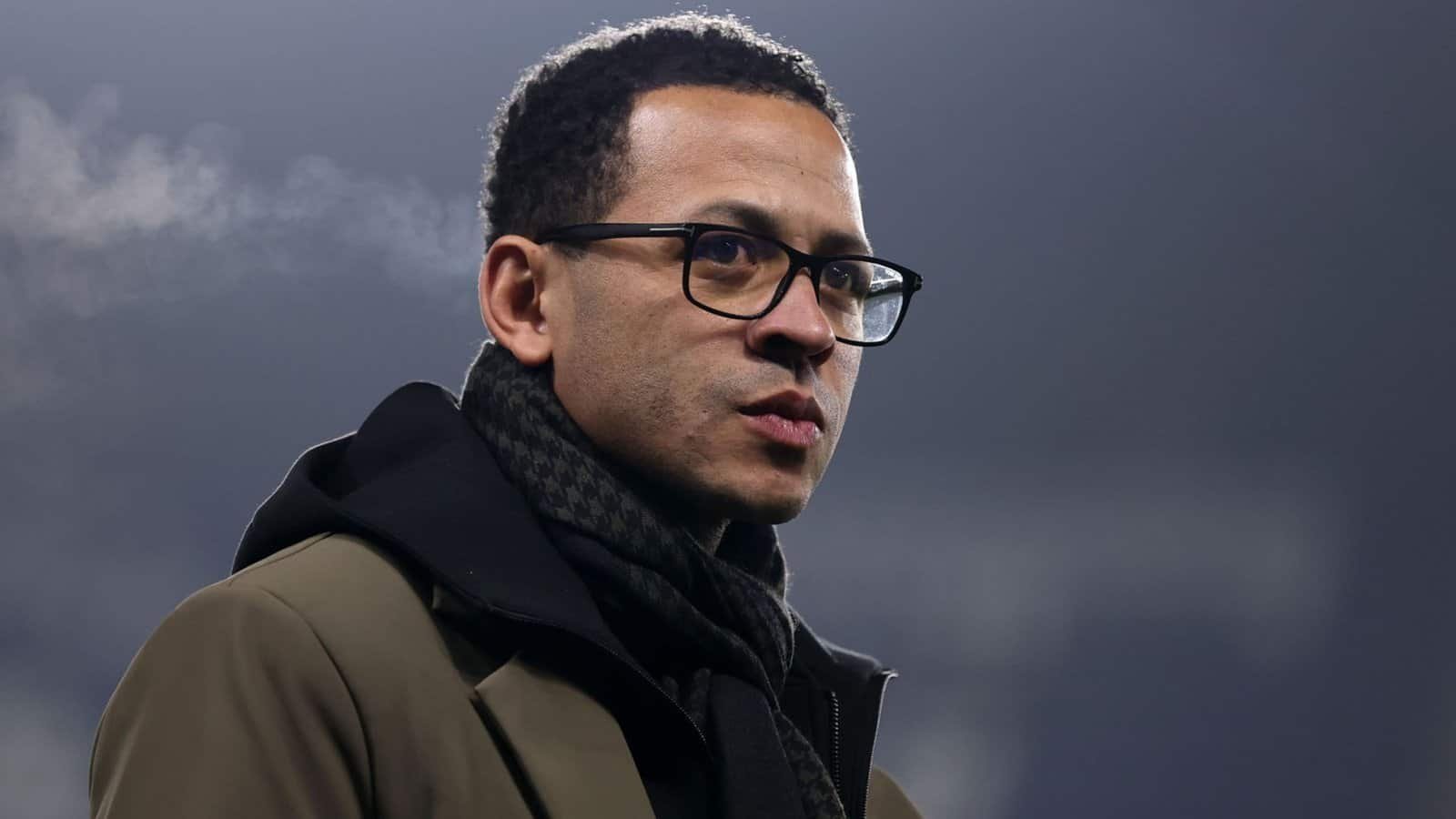 Liam Rosenior, vêtu d'un manteau sombre et portant des lunettes, regarde pensivement devant l'arrière-plan brumeux d'un stade, illustrant sa concentration sur le football.