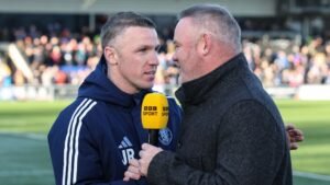 Wayne Rooney spreekt met een medecoach, beiden betrokken in een gesprek op het veld, met een menigte fans op de achtergrond.
