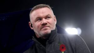 Wayne Rooney, vêtu d'un manteau sombre arborant un coquelicot, fixe un regard pensif sous les lumières du stade, méditant sur ses récentes décisions d'entraîneur.