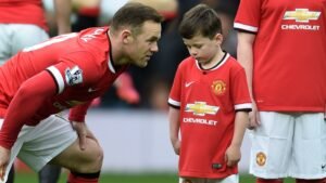 Wayne Rooney hurkt neer om met zijn zoon Kai te praten, beiden gekleed in het thuistenue van Manchester United, tijdens een wedstrijddag.