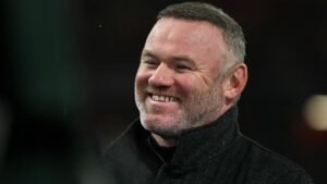 Wayne Rooney glimlacht warm terwijl hij een wedstrijd op Old Trafford bijwoont, gekleed in een zwarte jas tegen een wazig stadionachtergrond.