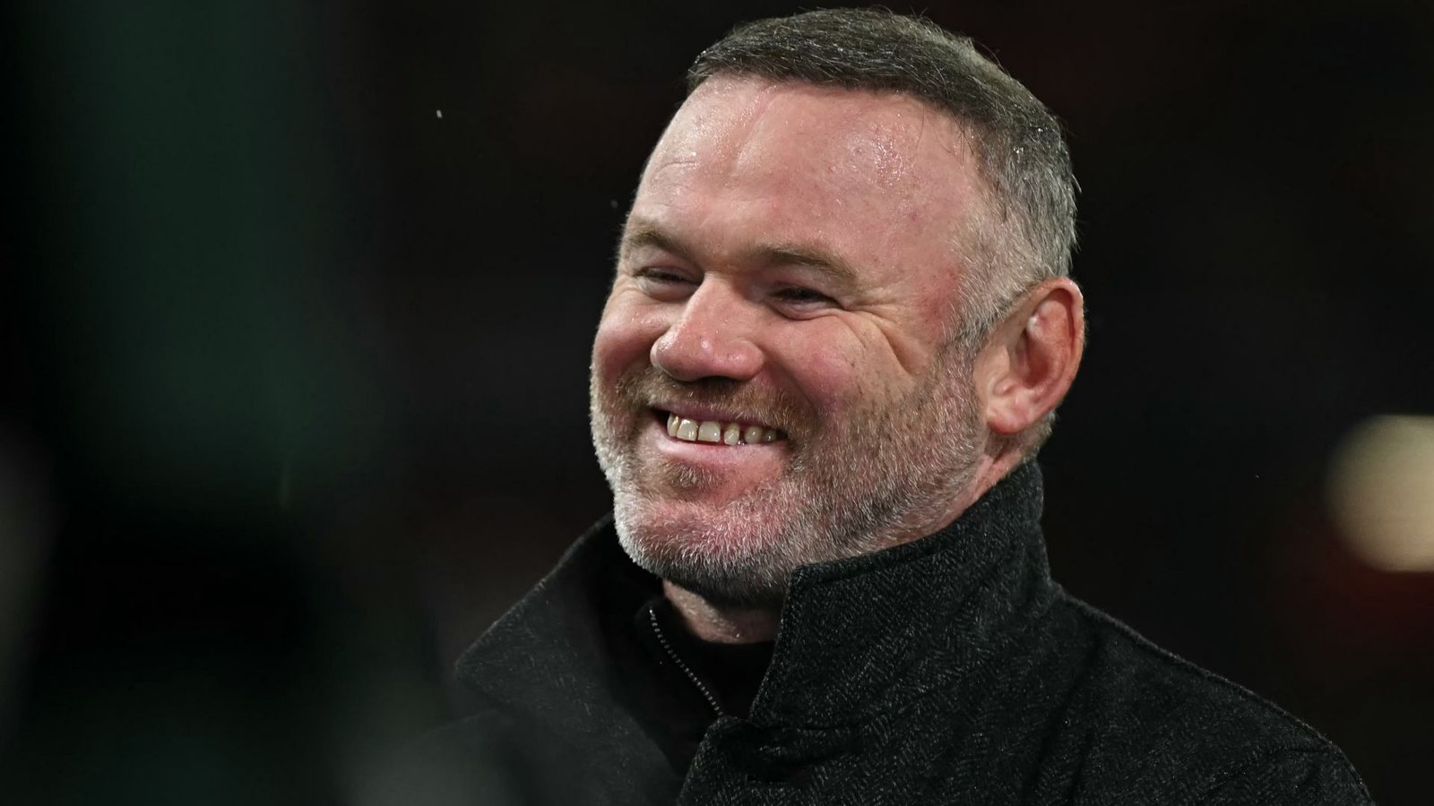 Wayne Rooney glimlacht warm terwijl hij een wedstrijd op Old Trafford bijwoont, gekleed in een zwarte jas tegen een wazig stadionachtergrond.