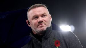 Wayne Rooney, vêtu d'un manteau sombre arborant un coquelicot, semble pensif sous les lumières du stade, laissant entrevoir son éventuel retour au coaching.