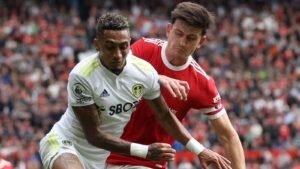 Le joueur de Leeds, en tenue blanche avec des accents jaunes, se confronte au défenseur de Manchester United, tous deux luttant pour le ballon au milieu d'une foule animée.