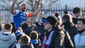 Macclesfield FC-kapitein viert met uitzinnige fans na een overwinning in de FA Cup, gekleed in een blauwe outfit met een oranje armband, temidden van een juichende menigte.