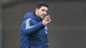 Mikel Arteta fait des gestes stratégiques pendant une séance d'entraînement d'Arsenal, portant une veste matelassée bleue arborant le logo du club.