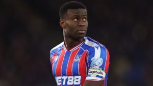 Marc Guehi, gekleed in het gestreepte rood-blauwe tenue van Crystal Palace, kijkt bedachtzaam opzij, waarbij zijn aanvoerdersband zichtbaar is.