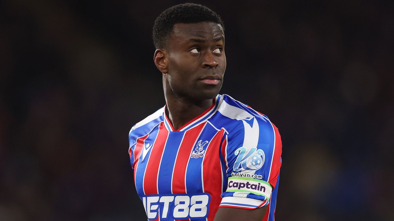 Marc Guehi, gekleed in het gestreepte rood-blauwe tenue van Crystal Palace, kijkt bedachtzaam opzij, waarbij zijn aanvoerdersband zichtbaar is.