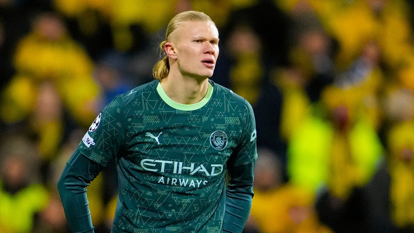 Erling Haaland in zijn groene Manchester City-tenue, geconcentreerd kijkend te midden van een menigte geelgeklede fans tijdens een wedstrijd.