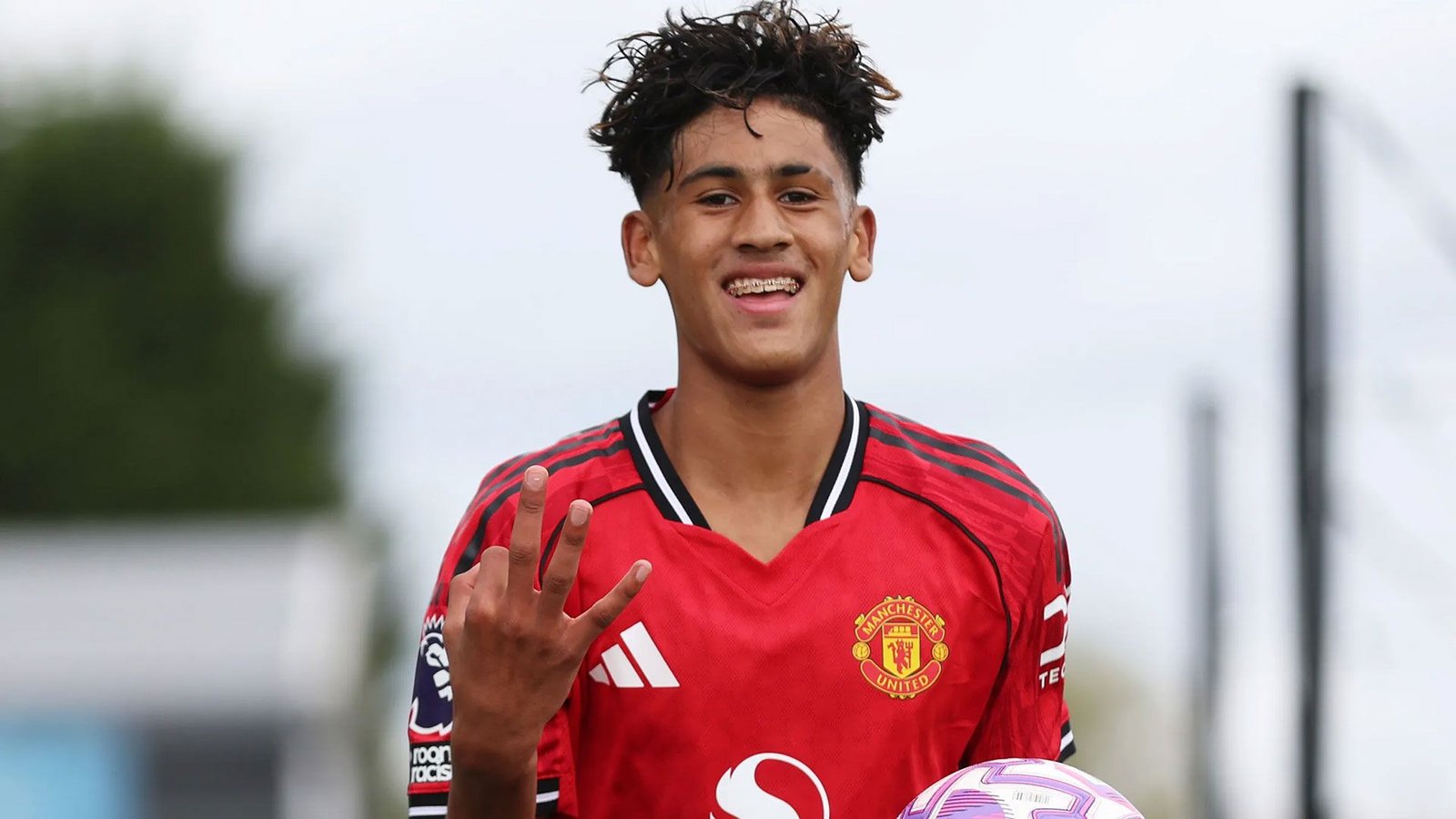 JJ Gabriel viert met een Manchester United-shirt, houdt een voetbal vast en toont vier vingers, tegen een bewolkte achtergrond.