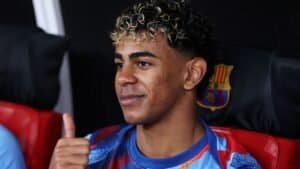 Young football player with curly hair and Moroccan jersey, smiling and giving a thumbs up, at a Moroccan sports event.