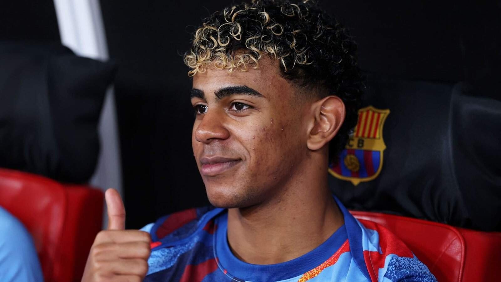 Young football player with curly hair and Moroccan jersey, smiling and giving a thumbs up, at a Moroccan sports event.