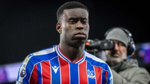 Marc Guéhi dans le maillot rouge et bleu de Crystal Palace, affichant une expression sérieuse, avec un caméraman en arrière-plan.