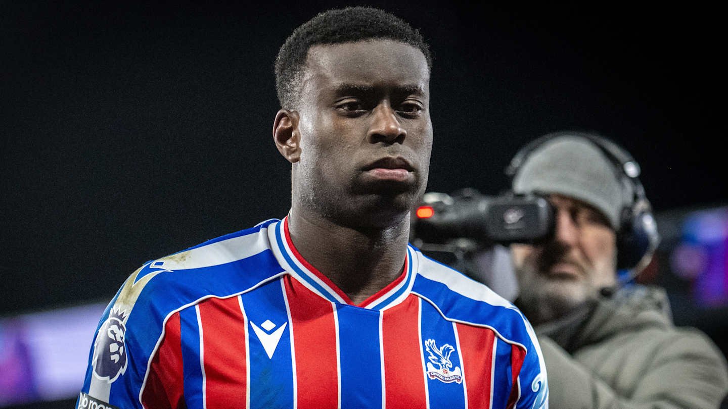 Marc Guéhi dans le maillot rouge et bleu de Crystal Palace, affichant une expression sérieuse, avec un caméraman en arrière-plan.