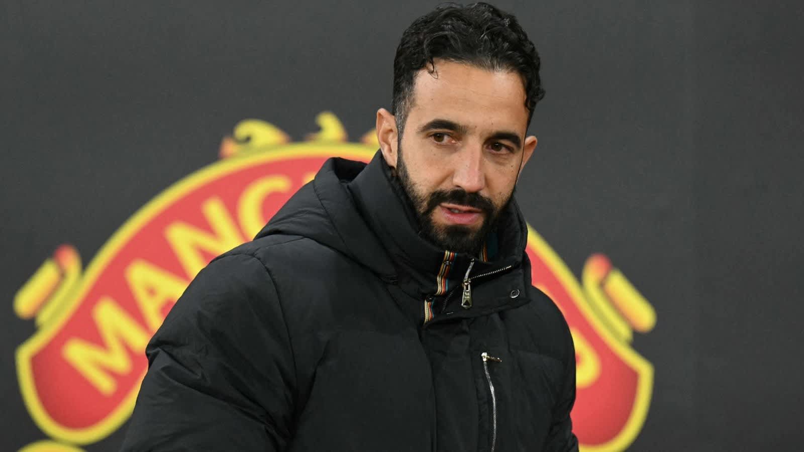 Rúben Amorim in een zwart jasje, peinzend voor een achtergrond met het logo van Manchester United.