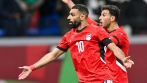Mohamed Salah celebrates a goal in Egypt's red kit, with a teammate cheering behind him during a crucial match.