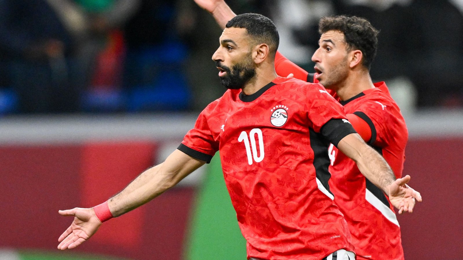 Mohamed Salah celebrates a goal in Egypt's red kit, with a teammate cheering behind him during a crucial match.