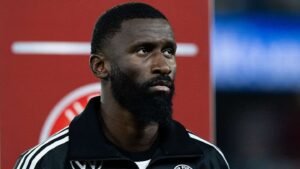 Antonio Rüdiger dans une veste d'entraînement noire, l'air sérieux devant un arrière-plan flou avec des accents rouges, réfléchissant à son avenir.