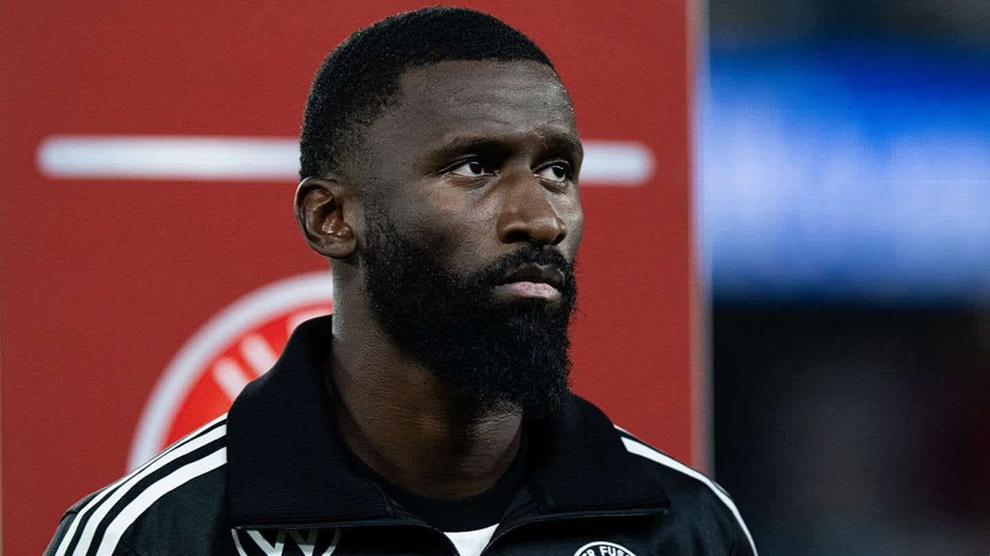 Antonio Rüdiger dans une veste d'entraînement noire, l'air sérieux devant un arrière-plan flou avec des accents rouges, réfléchissant à son avenir.