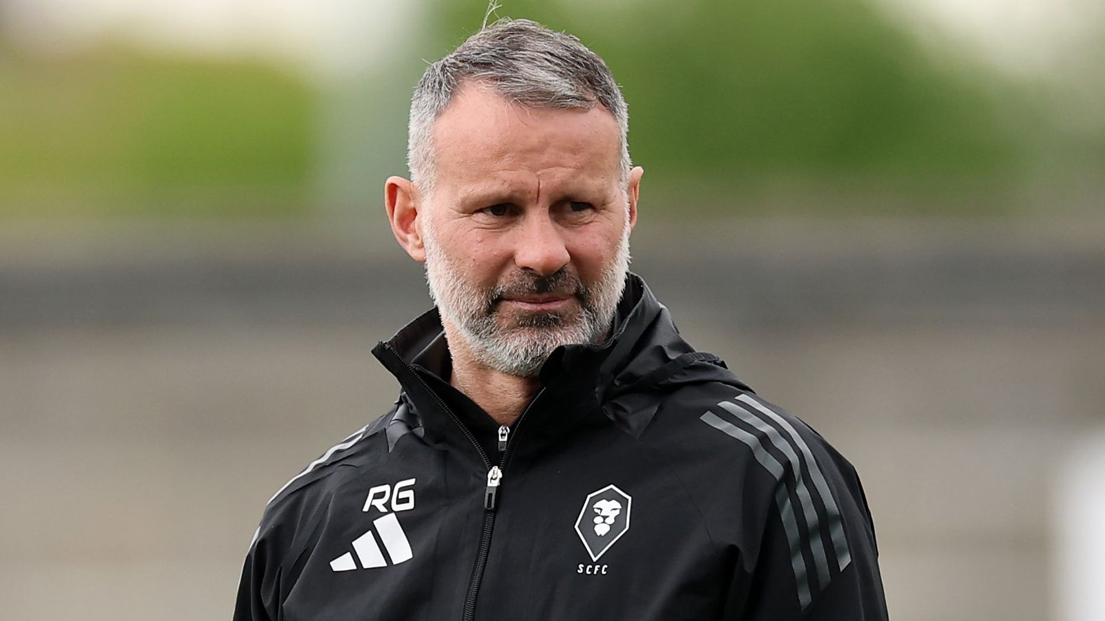 Ryan Giggs, gekleed in een zwart trainingsjack met initialen ‘RG’, lijkt in gedachten verzonken op het veld, wat zijn leiderschapsrol weerspiegelt.