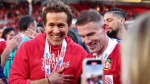 Ryan Reynolds et Jordan Davies célèbrent ensemble, vêtus de rouge, dans un stade bondé de supporters.