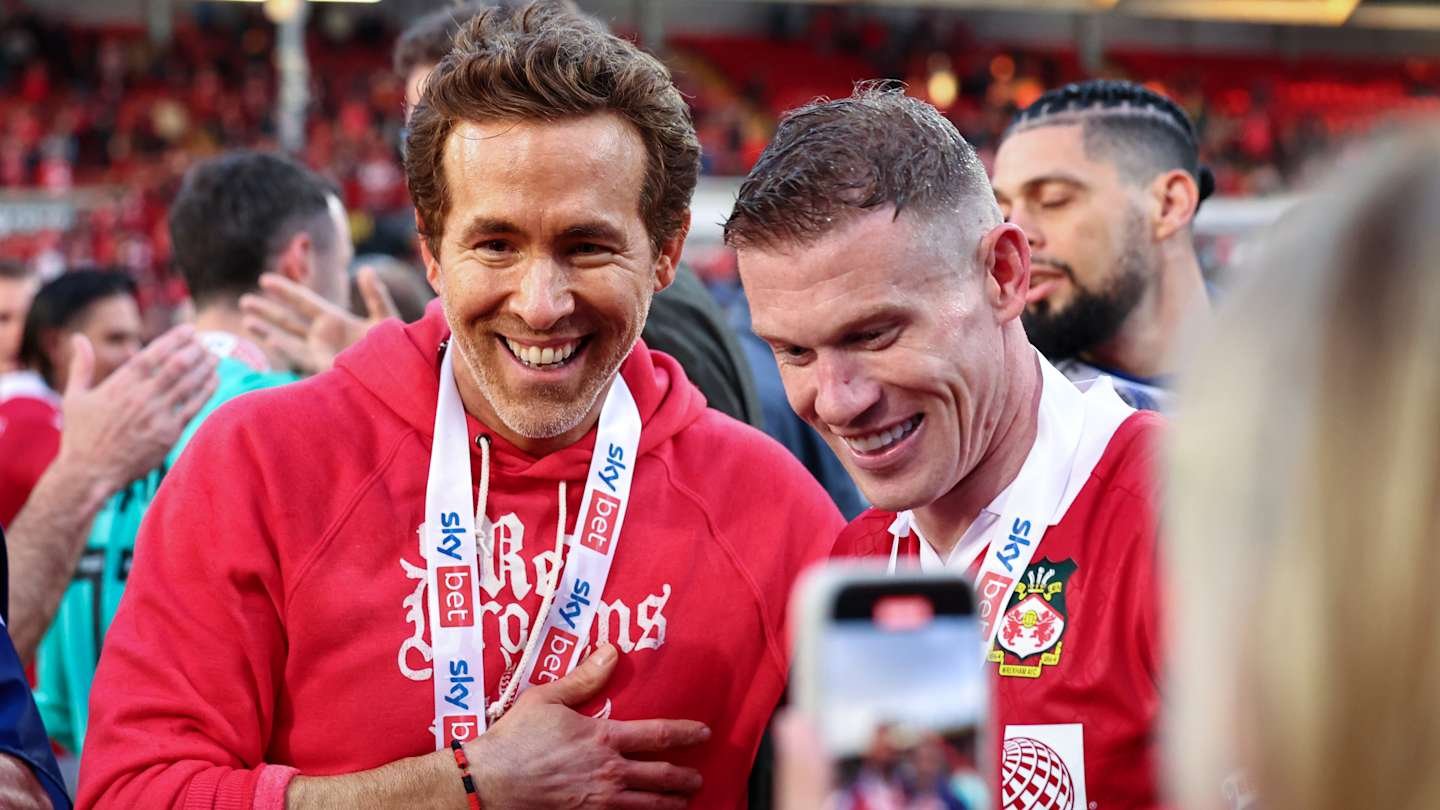 Ryan Reynolds et Jordan Davies célèbrent ensemble, vêtus de rouge, dans un stade bondé de supporters.
