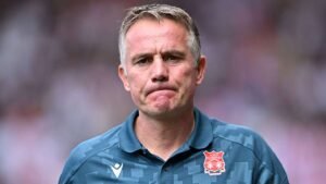 Le manager de Wrexham affiche une expression solennelle, portant un haut d'entraînement bleu-vert avec le logo du club, sur fond de stade flou.