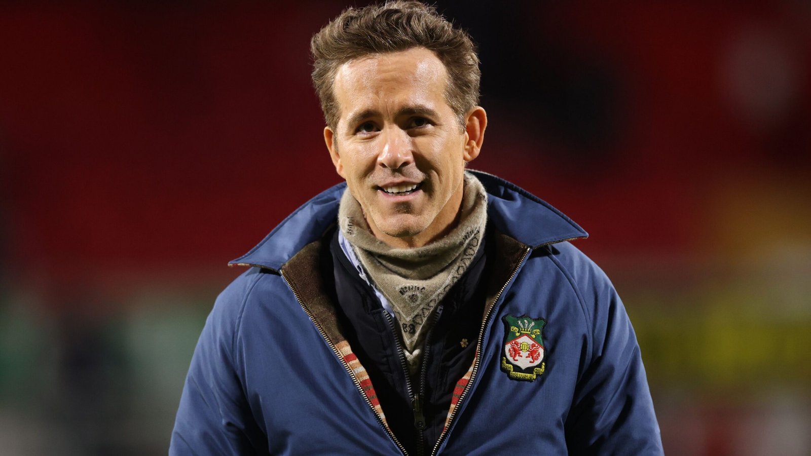 Ryan Reynolds smiles in a Wrexham jacket, showcasing team pride against a blurred stadium backdrop, capturing his engagement with football.