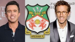 Ryan Reynolds en Rob McElhenney flankeren het Wrexham FC-clublogo, wat hun eigendom en betrokkenheid bij de wederopstanding van de club benadrukt.