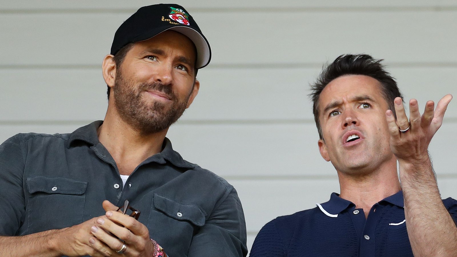 Ryan Reynolds en Rob McElhenney waren in gesprek verwikkeld in het stadion van Wrexham, waarbij Reynolds een zwarte pet droeg en McElhenney levendig gebaren maakte.