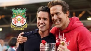 Rob McElhenney et Ryan Reynolds célèbrent avec un trophée au Wrexham FC, affichant des sourires devant l'arrière-plan vibrant du stade.