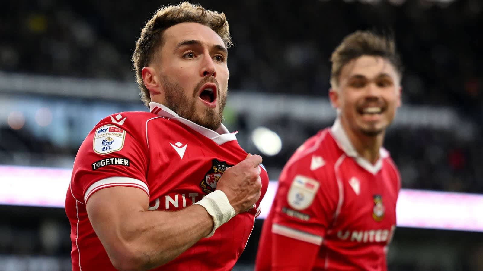 Les joueurs de Wrexham célèbrent après un but, l'un d'eux enlevant son maillot sous l'effet de l'excitation, portant un équipement rouge avec des détails blancs.