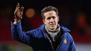 Ryan Reynolds waves to fans, dressed in a blue coat with a Wrexham logo, showcasing a warm, friendly gesture during a match.