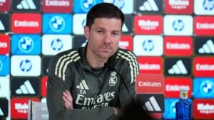 Xabi Alonso est assis à une table de conférence de presse, portant un survêtement du Real Madrid, avec des logos de marque en arrière-plan coloré.