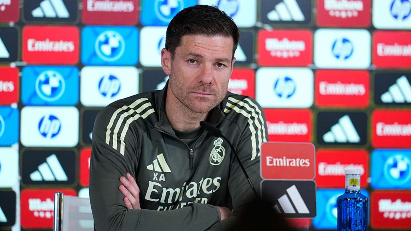 Xabi Alonso est assis à une table de conférence de presse, portant un survêtement du Real Madrid, avec des logos de marque en arrière-plan coloré.