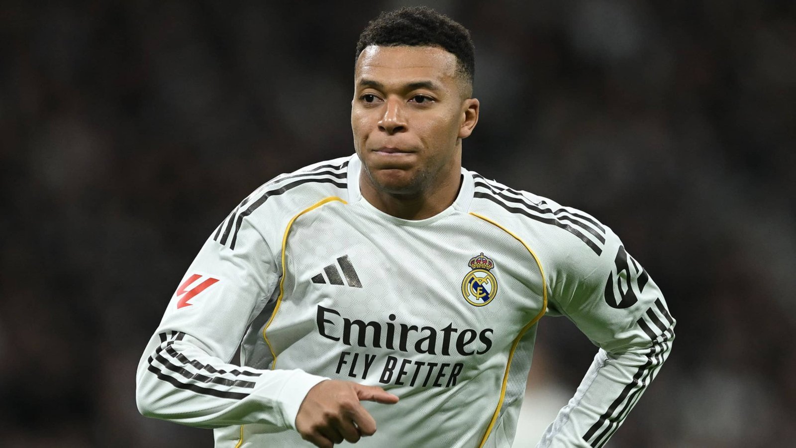 كيليان مبابي يرتدي قميص ريال مدريد الأبيض، مركزاً على أرض الملعب خلال مباراة، مع إبراز شعار النادي والراعي.