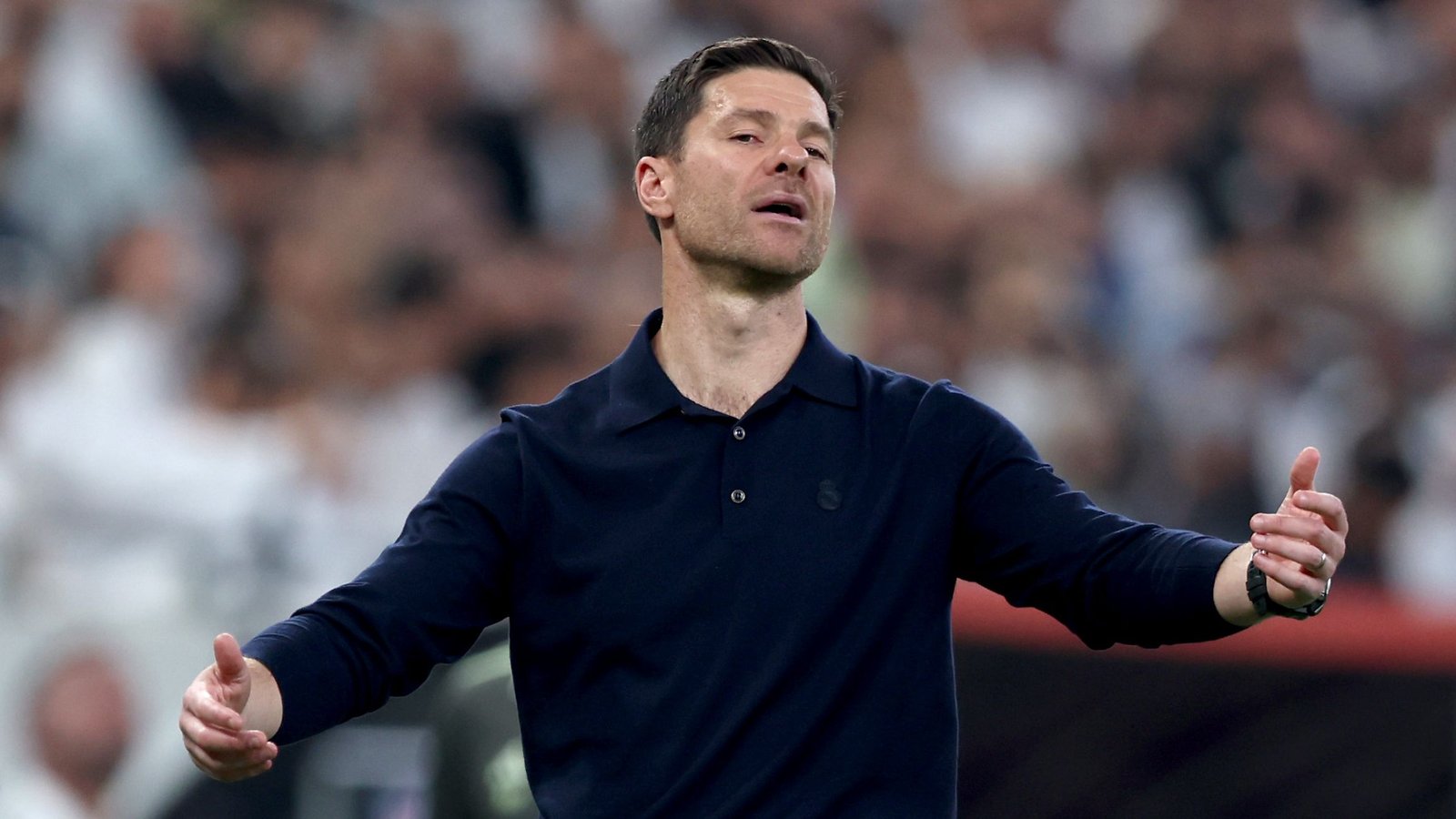 Xabi Alonso gebaart expressief aan de zijlijn, gekleed in een donker poloshirt met het logo van Real Madrid, tegen een wazige achtergrond van toeschouwers.