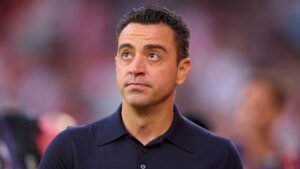 Xavi, in een marineblauwe polo, kijkt nadenkend omhoog met een vervaagde stadionachtergrond, te midden van speculaties over zijn toekomst in het coachen.
