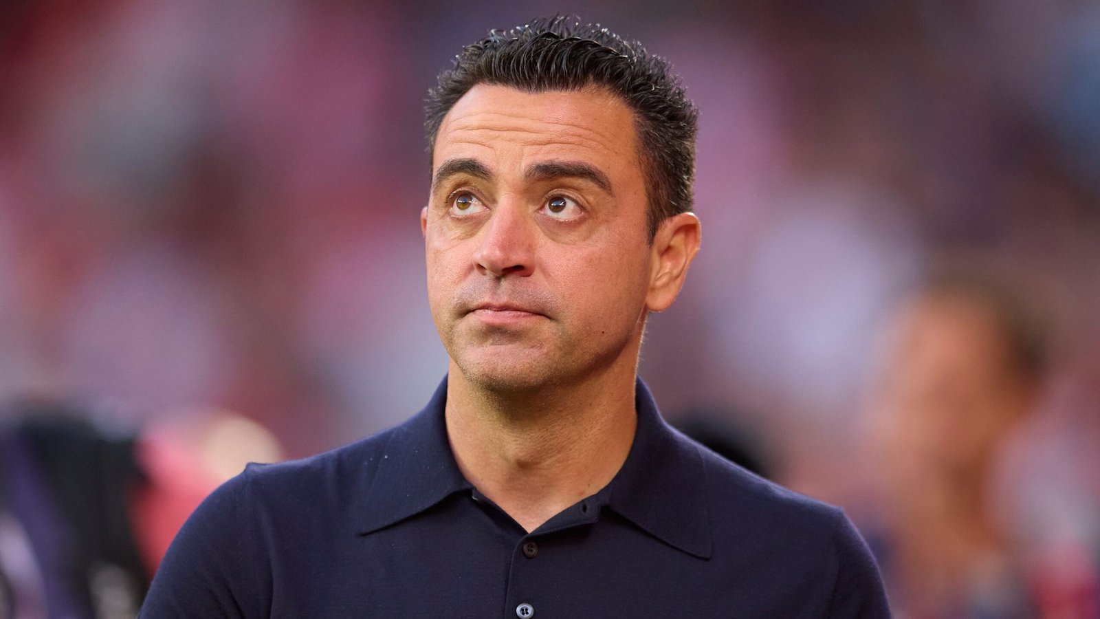 Xavi, in een marineblauwe polo, kijkt nadenkend omhoog met een vervaagde stadionachtergrond, te midden van speculaties over zijn toekomst in het coachen.