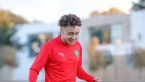 Moroccan footballer in a red Puma training kit smiles during a training session, with blurred buildings in the background.