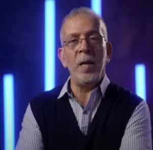 Analyste sportif marocain parlant, portant des lunettes et un gilet sombre, avec un fond de lumières néon bleues.