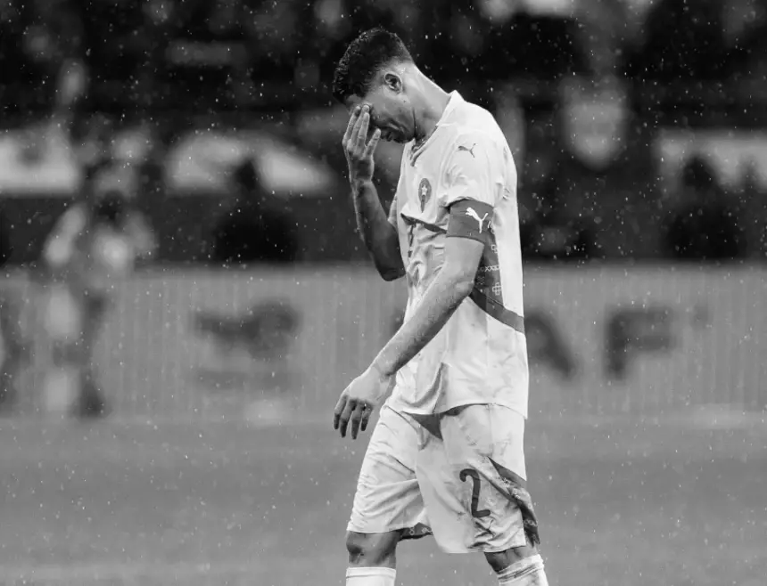 لاعب مغربي يظهر إحباطاً خلال مباراة ممطرة، مرتدياً قميص المنتخب الوطني الأبيض بتفاصيل حمراء.