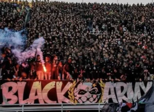 Dense crowd of passionate fans in black attire, cheering with flares, displaying a large "BLACK ARMY" banner at a football match.