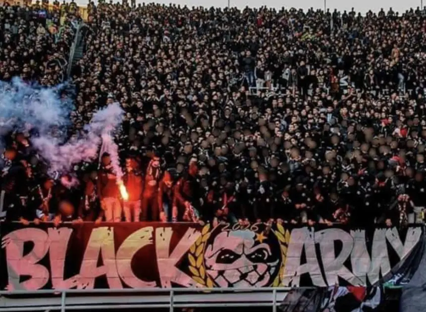 Dense crowd of passionate fans in black attire, cheering with flares, displaying a large "BLACK ARMY" banner at a football match.