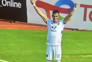 Marokkaanse voetballer viert een doelpunt met opgeheven armen, in een wit tenue met blauwe accenten, op een levendig groen veld.