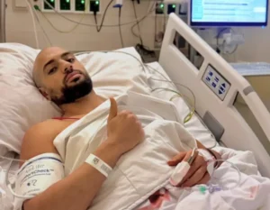 Moroccan footballer giving a thumbs up from a hospital bed, showcasing a determined expression despite medical equipment in the background.