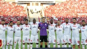 Wydad Casablanca-spelers poseren in hun witte tenues voor een wedstrijd, met een levendig publiek in rood achter hen, wat team eenheid uitstraalt.