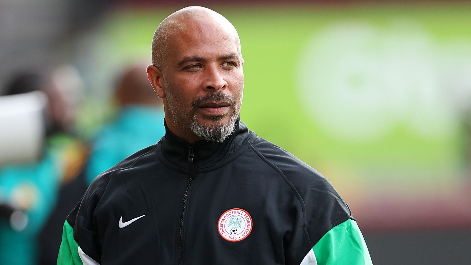 Assistant coach, wearing a black Nigeria Football Federation tracksuit, with a grey beard and focused expression, stands against a blurred green background.