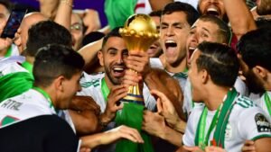 Les joueurs algériens célèbrent joyeusement, brandissant le trophée de la Coupe d'Afrique des Nations, vêtus de tenues vert et blanc avec des médailles autour du cou.
